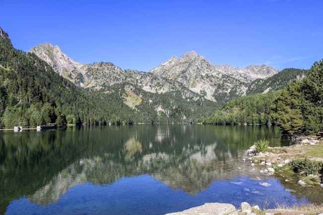 Lake with clear water surrounded by pine-clad hills and grey rocky mountains in Catalonia