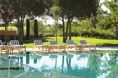 Outdoor swimming pool with greenery and trees in background and blue and white striped deck chairs around the pool edge
