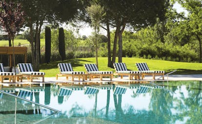 Outdoor swimming pool with greenery and trees in background and blue and white striped deck chairs around the pool edge