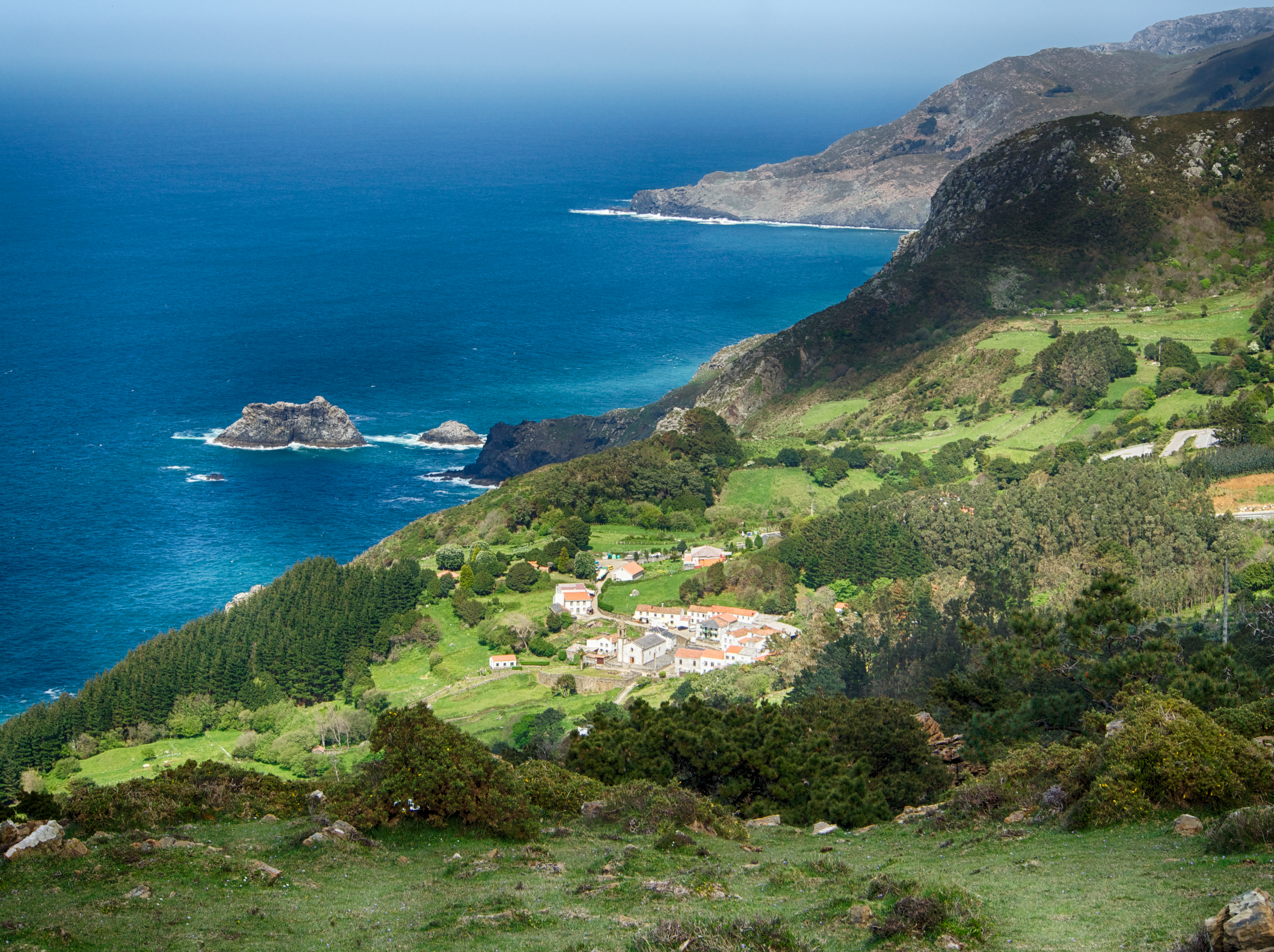 Coastline with green fields and hills dropping into turquoise sea with small village on flat field in Galicia