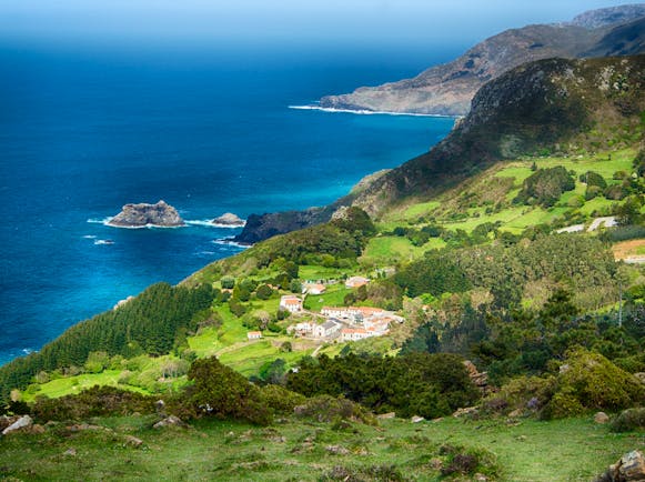 Coastline with green fields and hills dropping into turquoise sea with small village on flat field in Galicia