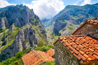 Rugged grey peaks with clouds and stone huts with red tiled roofs in foreground in the Picos de Europa