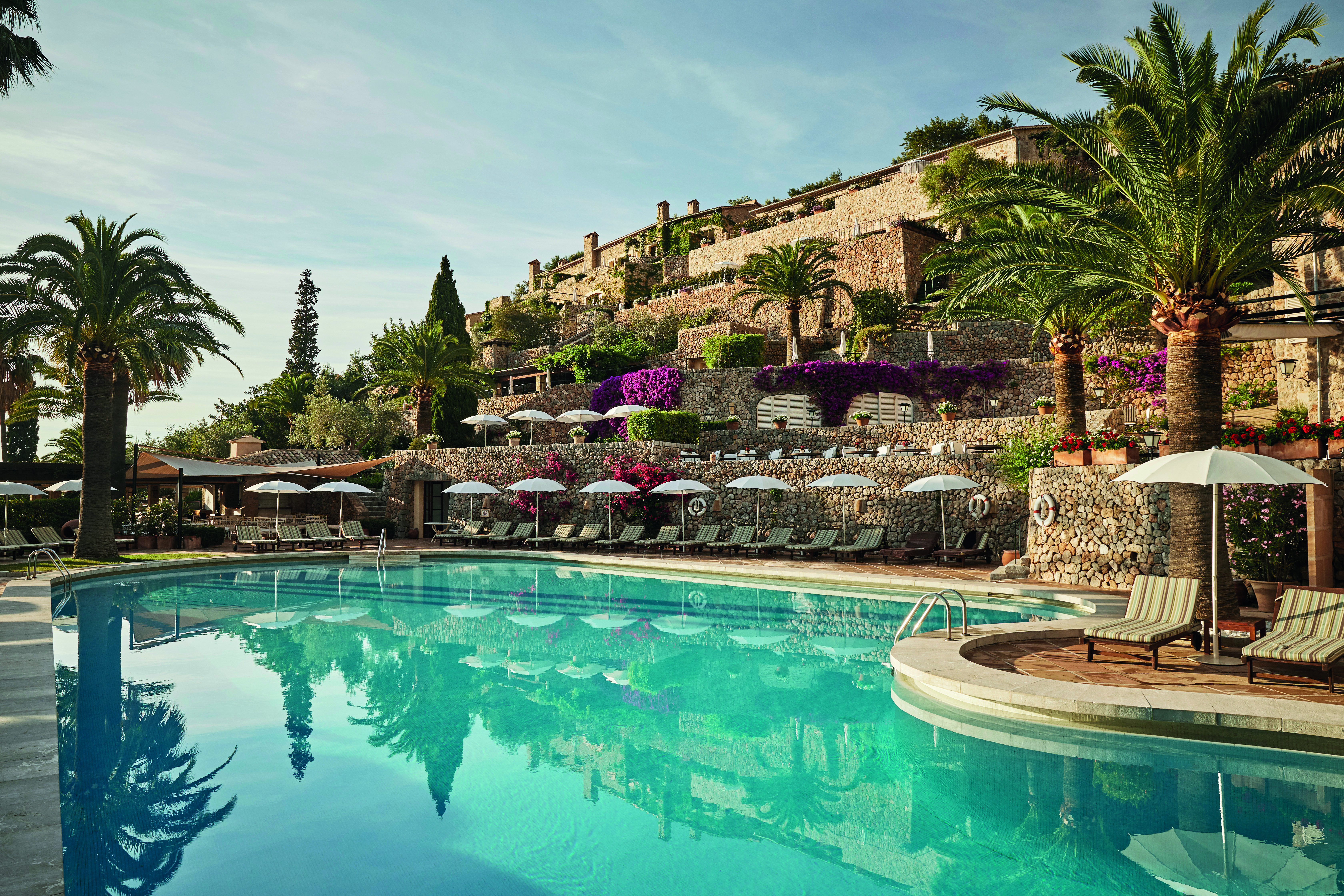 La Residencia Mallorca outdoor pool loungers umbrellas palm trees overlooked by stone buildings