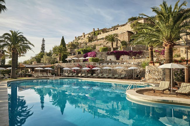 La Residencia Mallorca outdoor pool loungers umbrellas palm trees overlooked by stone buildings