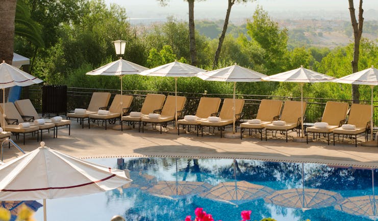 Outdoor pool with umbrellas and sun loungers set up around the pool and pink flowers shown nearby