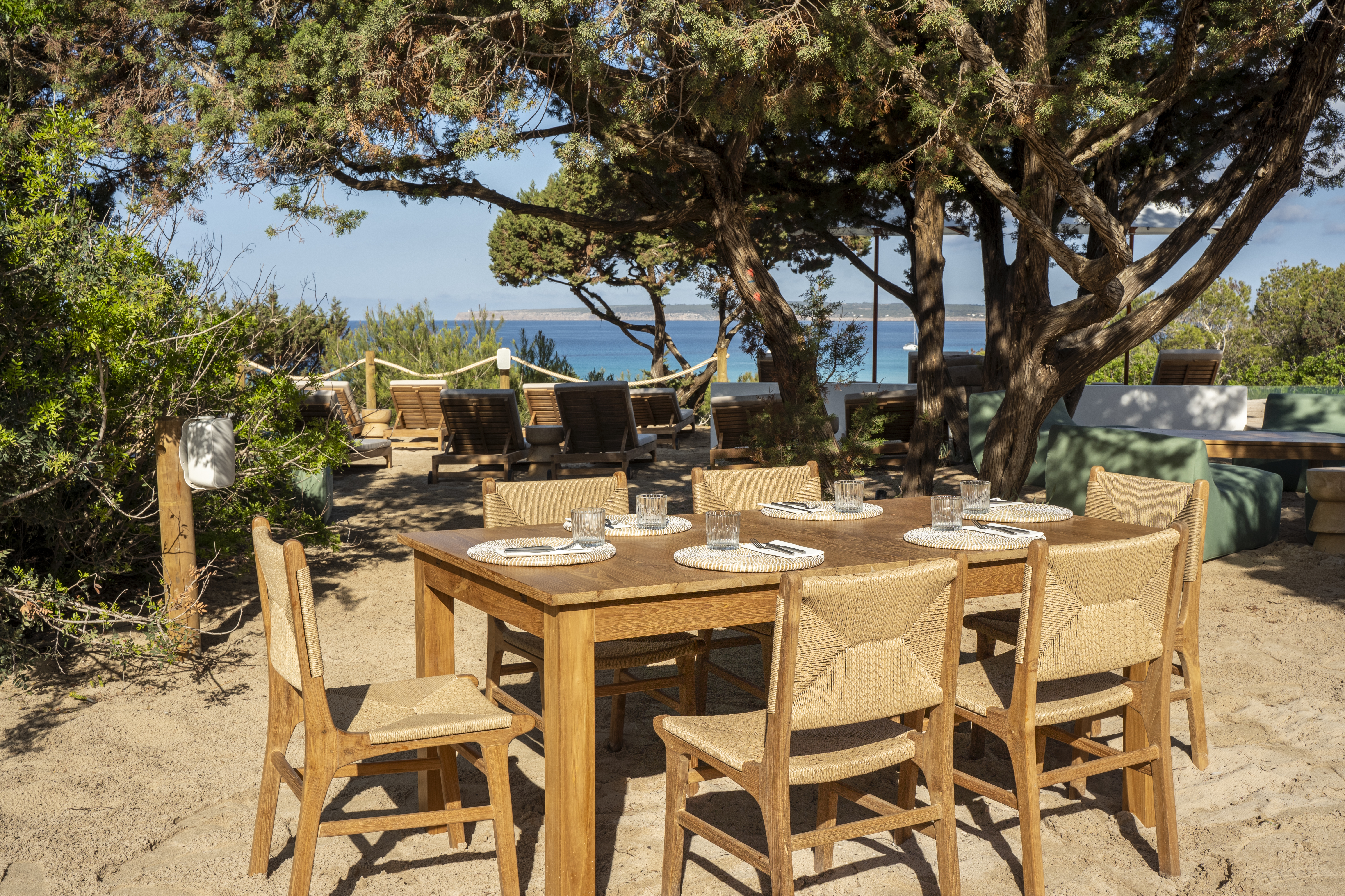 Dunas de Formentera dining with rustic wooden chairs and tables on the sand, with trees and the sea beyond