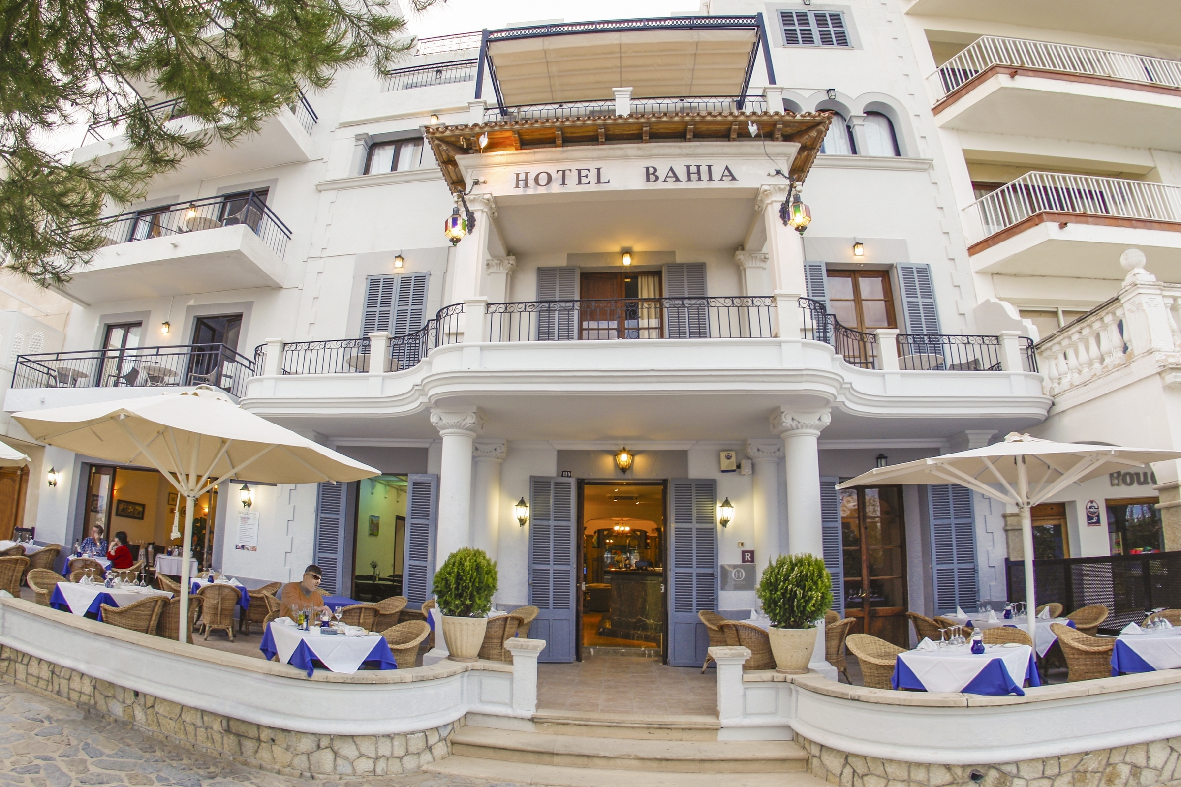 Hotel Hoposa Bahia entrance, showing the white walls and blue shutters, the restaurant terrace with blue clothed tables, and the balconies of rooms above
