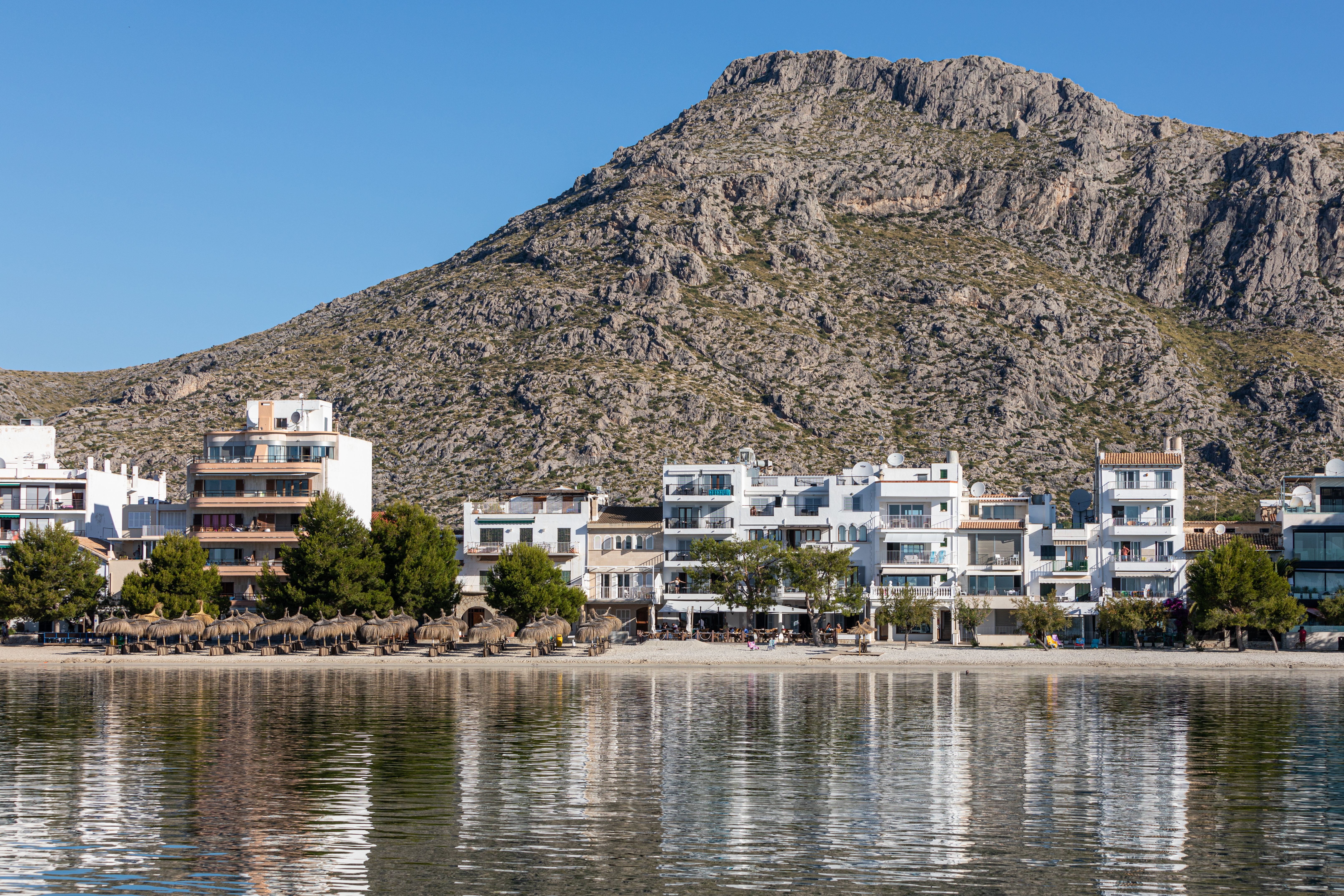 Hotel Hoposa Bahia seen from the sea looking back towards the sandy beach, the pine trees, the white hotel building, and the mountains behind