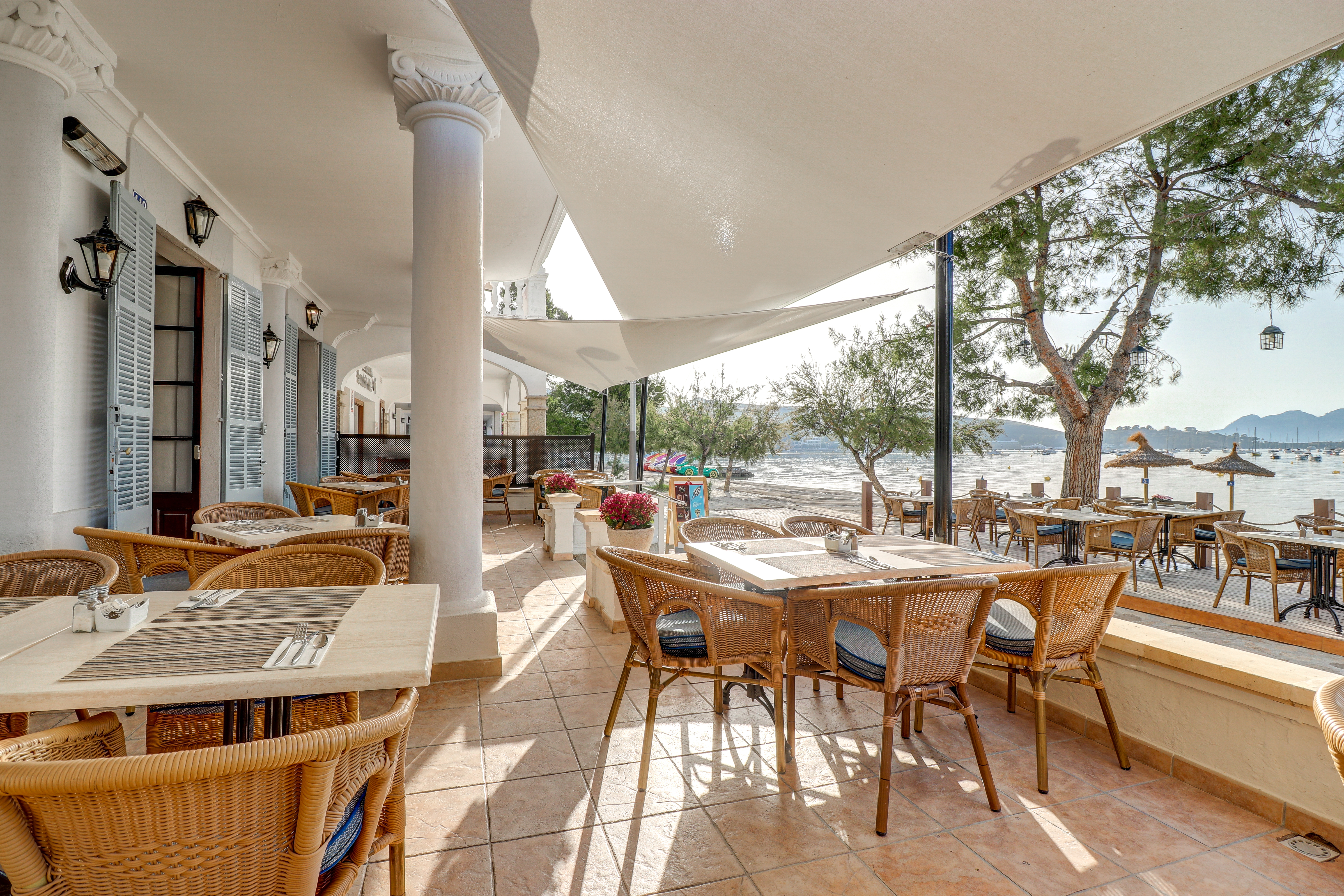 Hotel Hoposa Bahia restaurant terrace with terracotta tiles, blue and white tables and chairs, and pine trees and the sea visible beyond