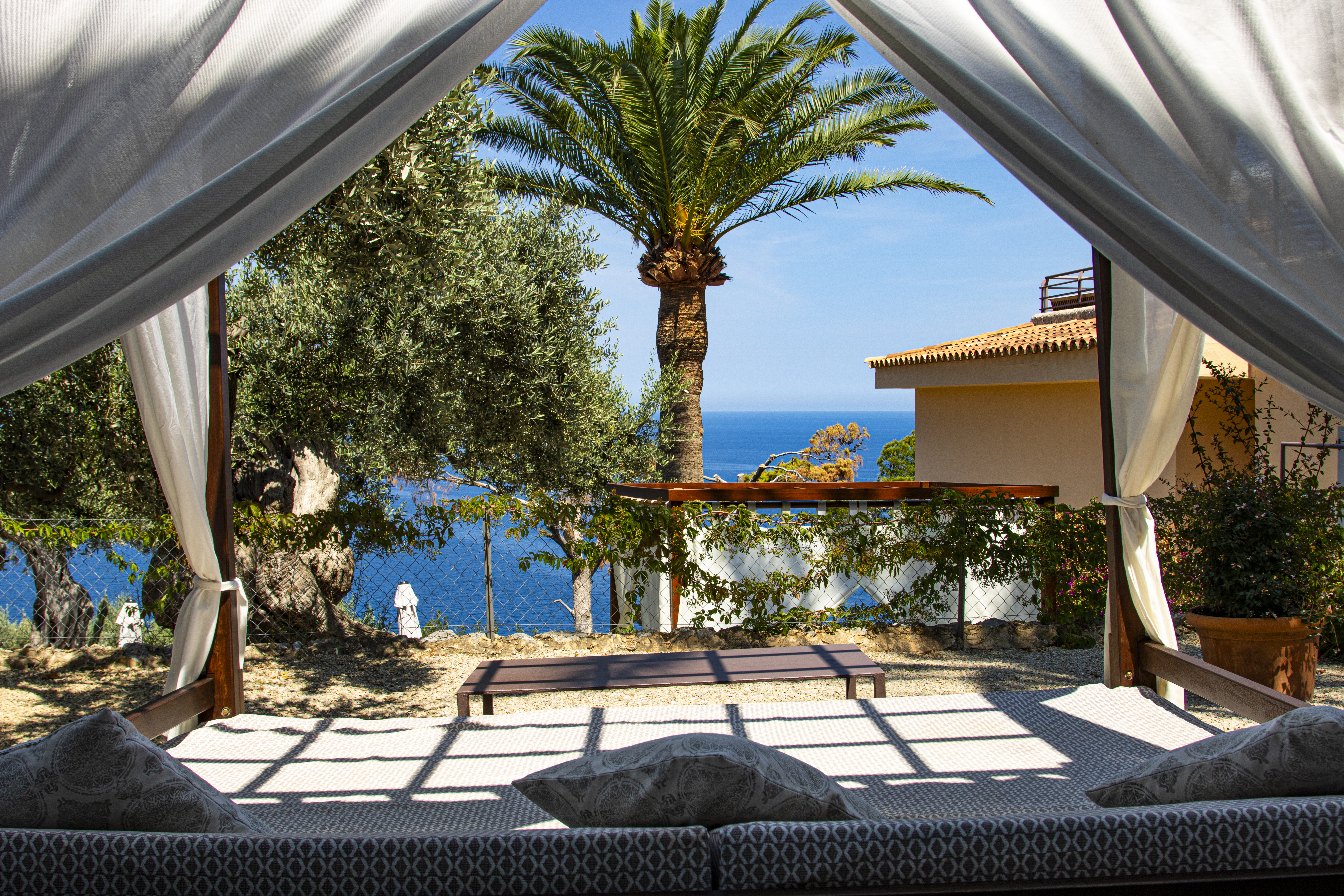 Hotel Hoposa Costa d'Or view out from beneath the canopy of a Balinese bed, looking across the hotel terrace, palm trees, and the sea in the distance