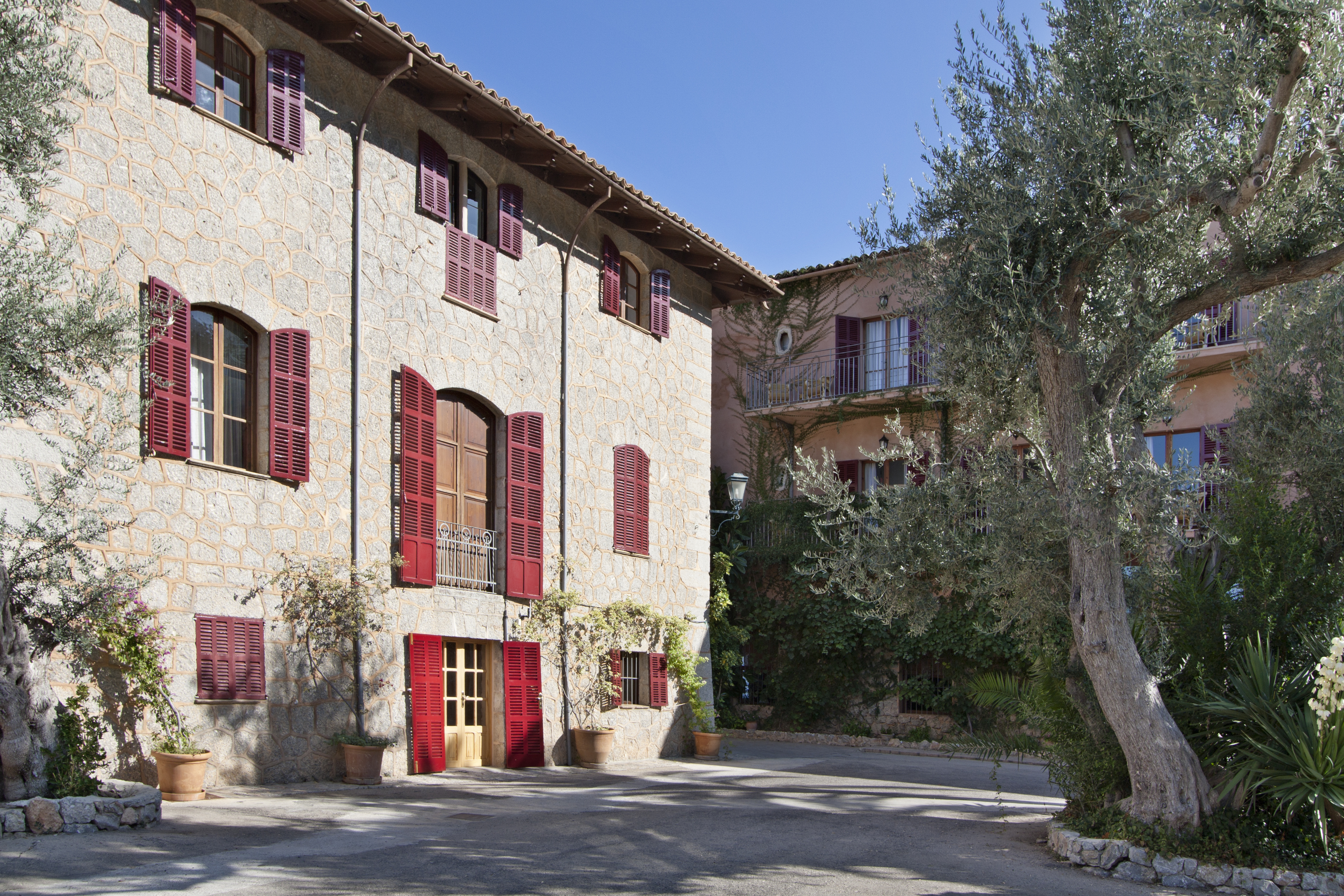 Hotel Hoposa Costa d'Or stone exterior with red-shuttered windows, terracotta plant pots beside the door, and a cobbled driveway complete with Aleppo pine trees