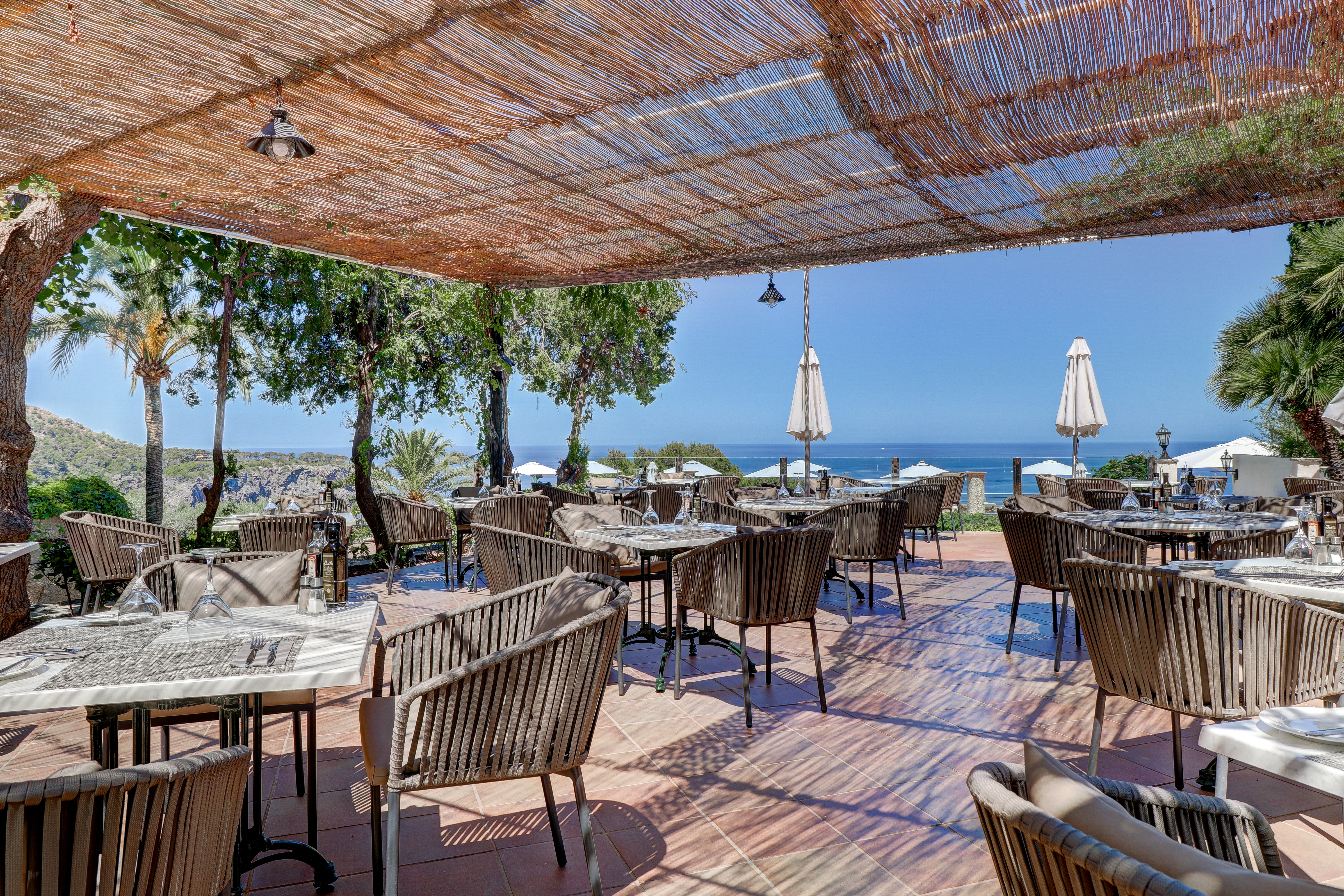 Hotel Hoposa Costa d'Or restaurant terrace, with rattan canopy over wooden tables and chairs, with trees and the sea visible beyond