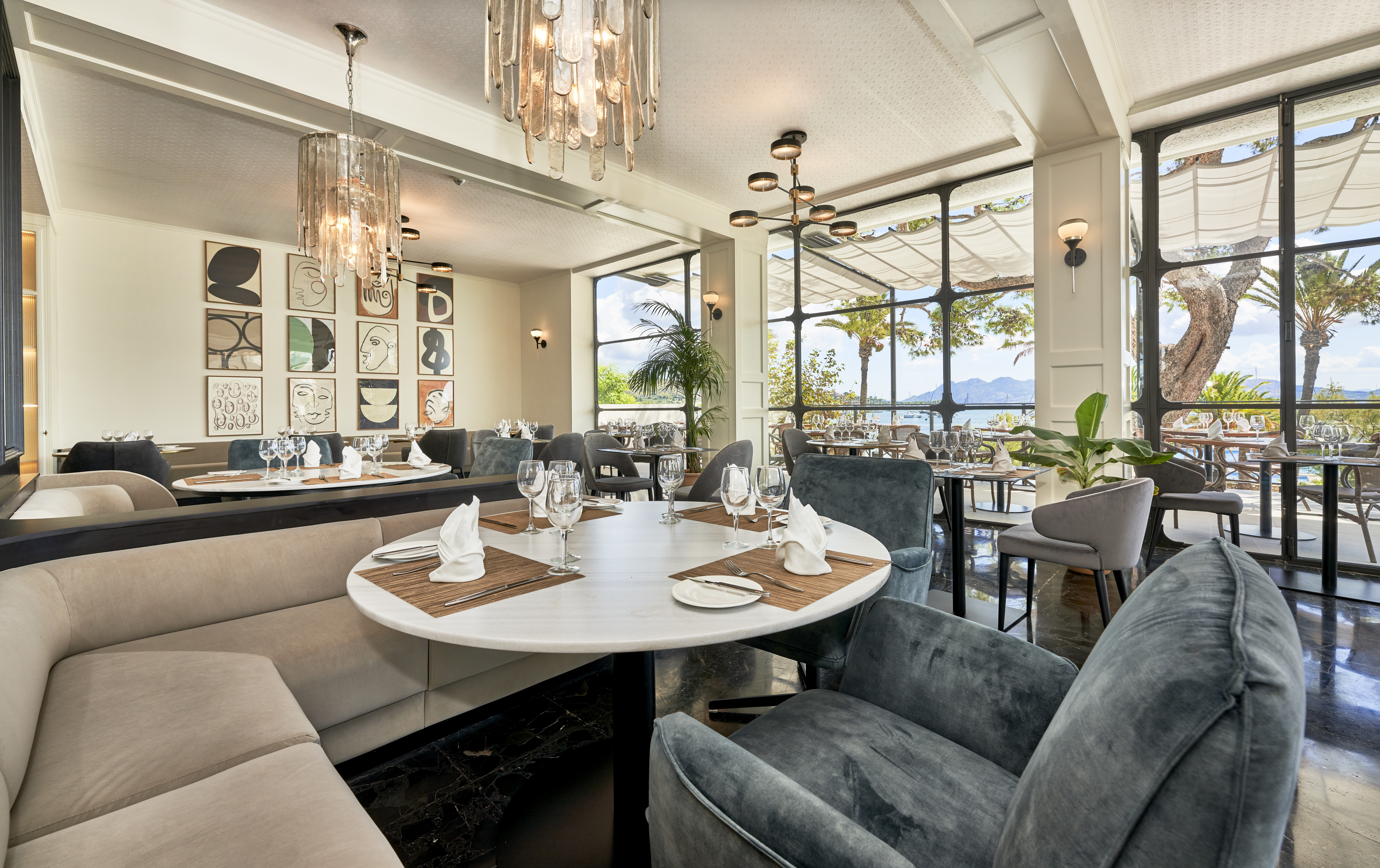 Hotel Hoposa Daina restaurant, with comfortable booth-style seats and armchairs, round tables, and floor-to-ceiling windows looking out towards the sea