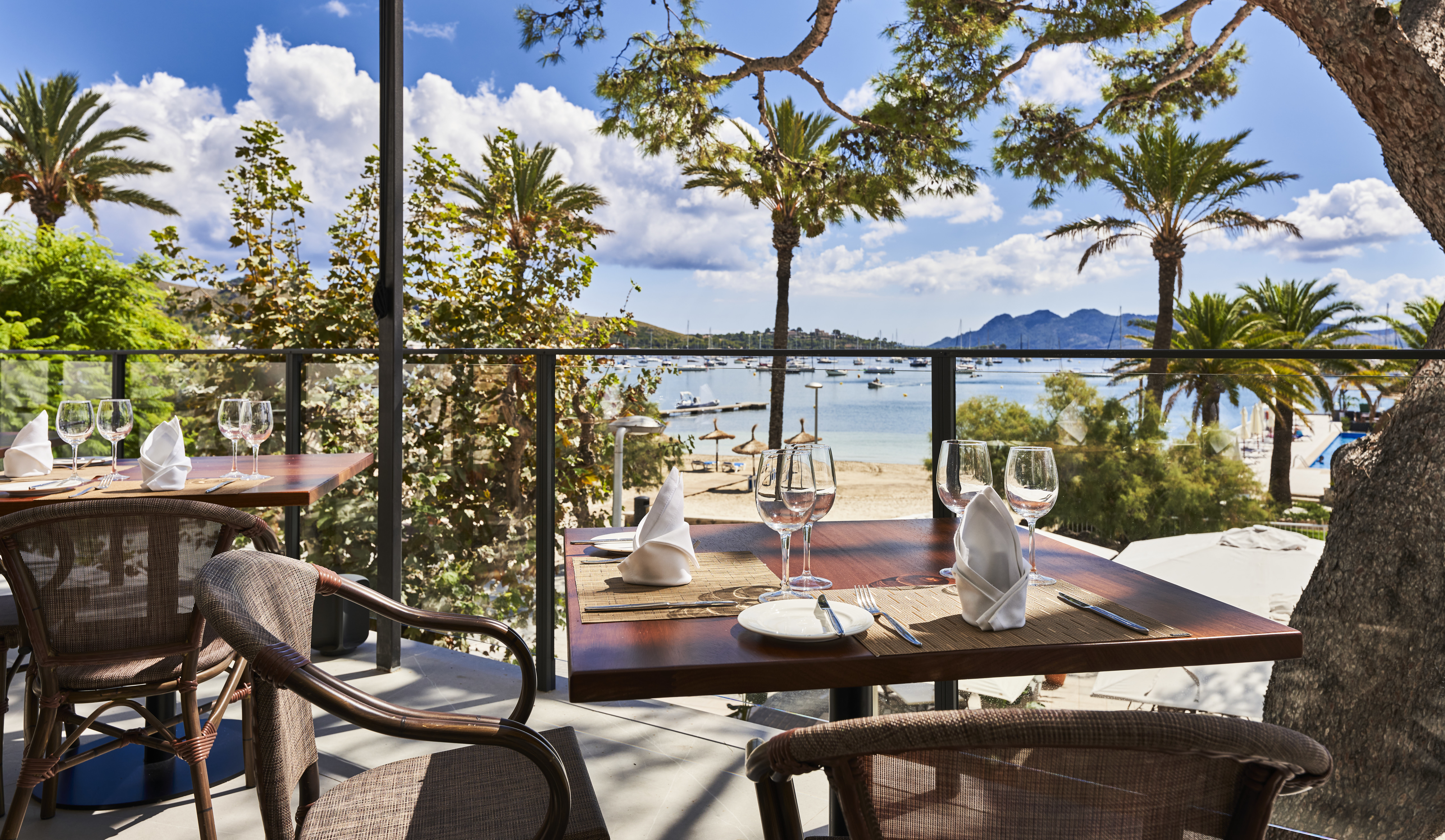 Hotel Hoposa Daina restaurant terrace with wooden tables and chairs beneath shady trees and a bright blue sky, with the sea visible beyond