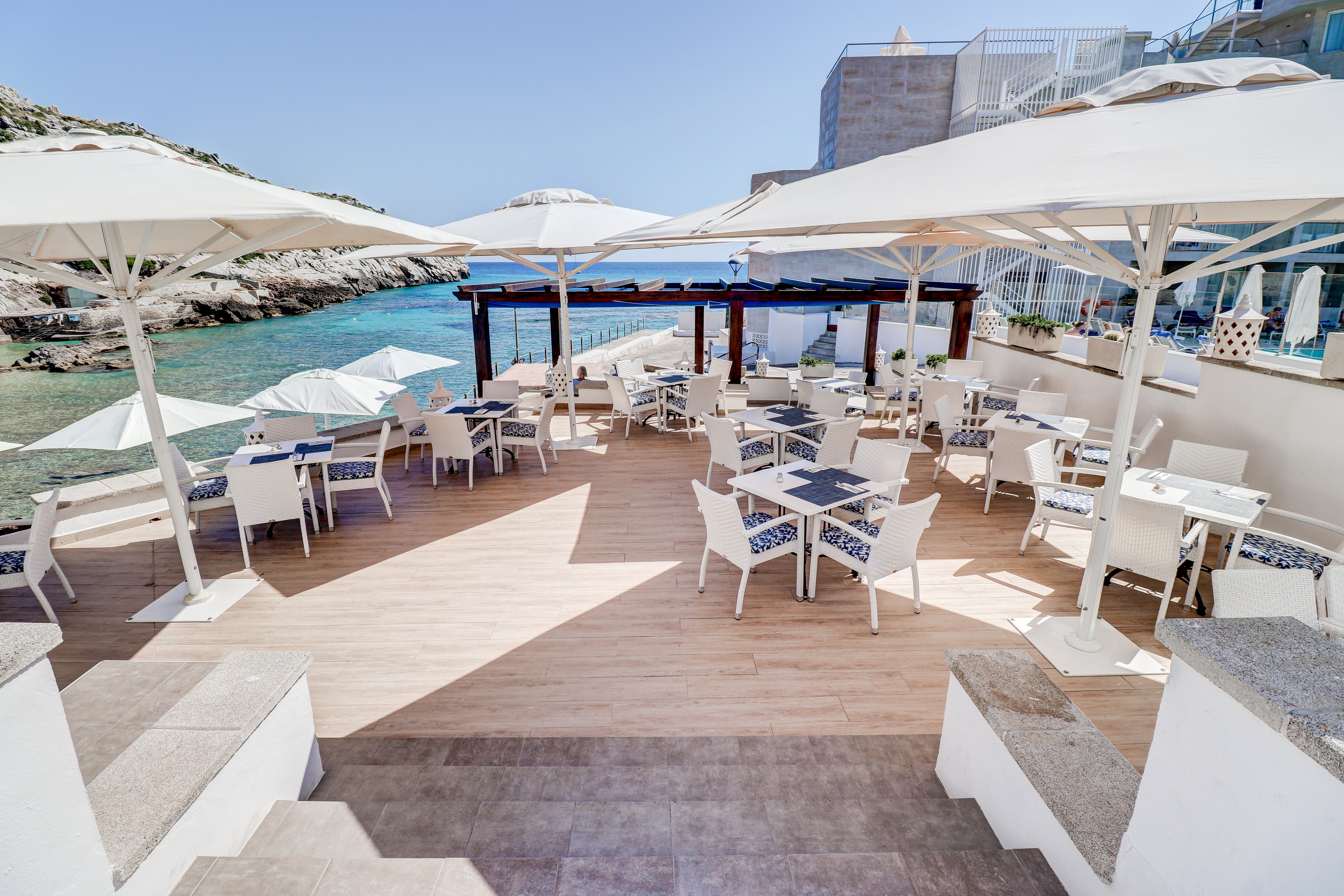 Hotel Hoposa Niu dining terrace, viewed from at the top of a short stairway, looking down on white tables and chairs, with the sea in the distance
