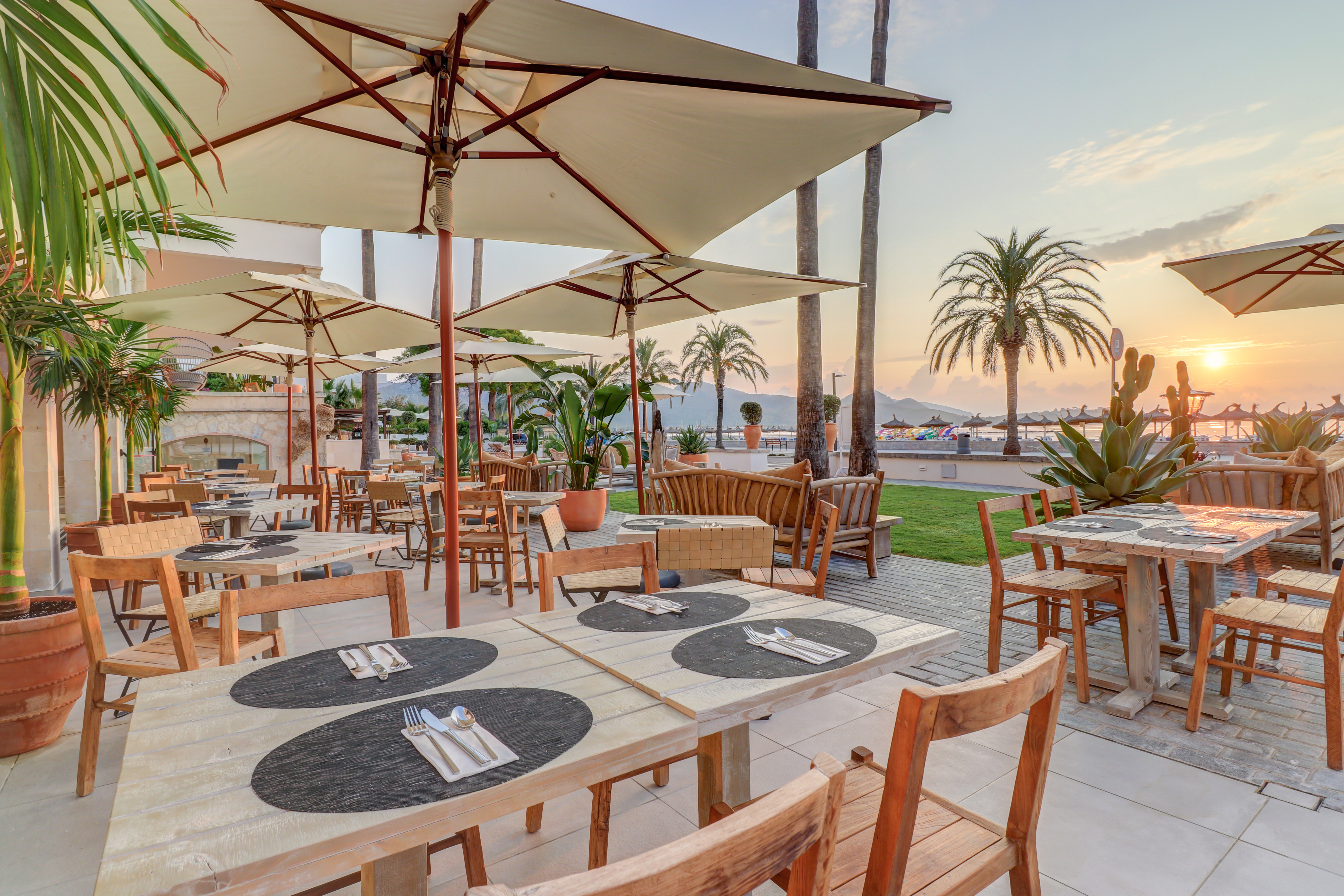 Hotel Hoposa Pollentia restaurant terrace in the evening, with simple wooden tables under square parasols, with the sea visible in the background