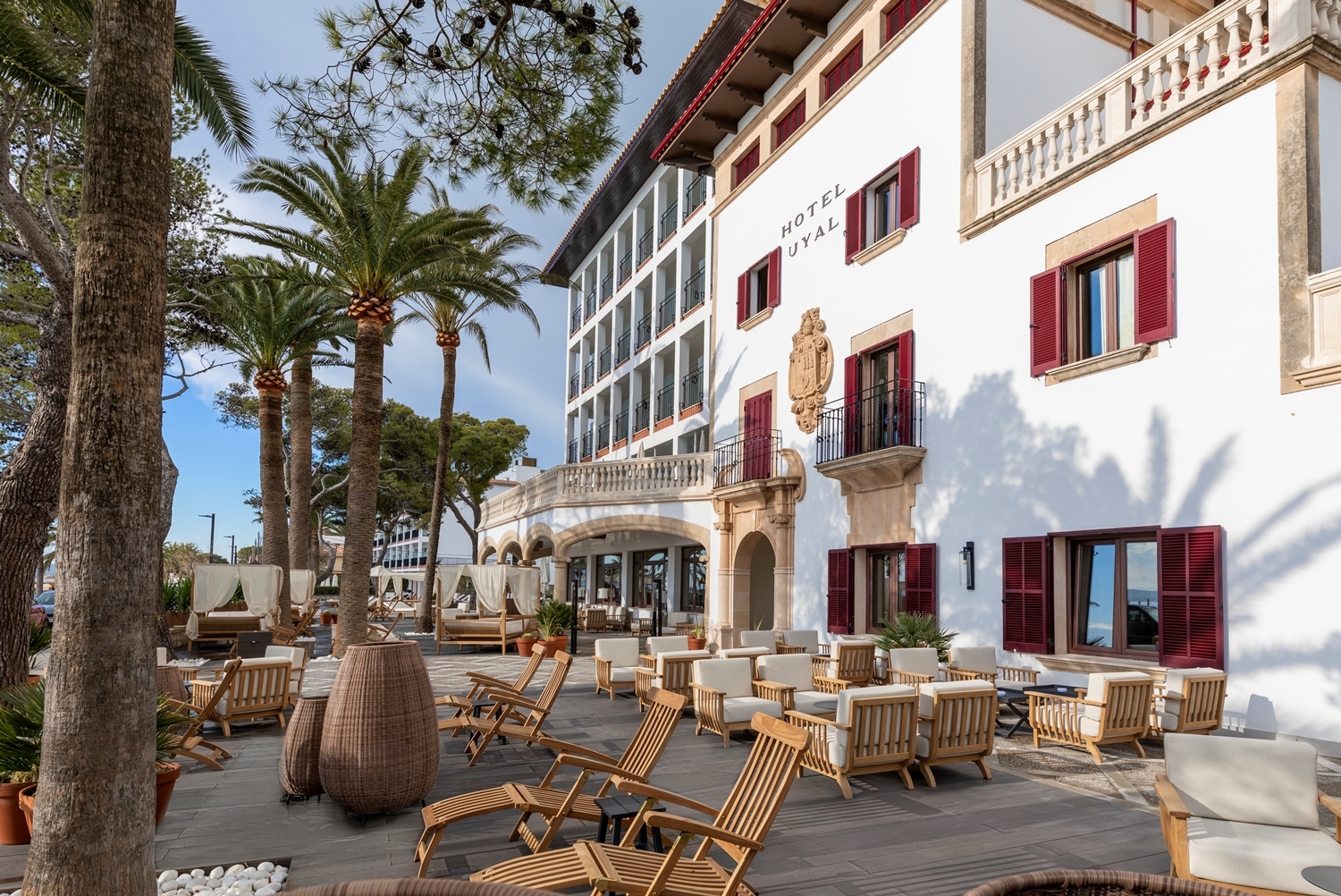 Hotel Hoposa Uyal ground floor terrace, with wooden loungers, comfortable sofas, and palm trees in front of the white hotel building with its red window shutters and modern glass extension