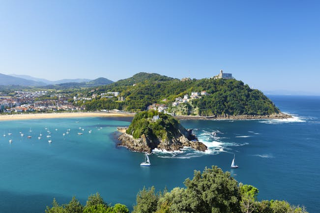 Sailing boats on blue sea in front of small island in bay with yellow sand beach in background at San Sebastian