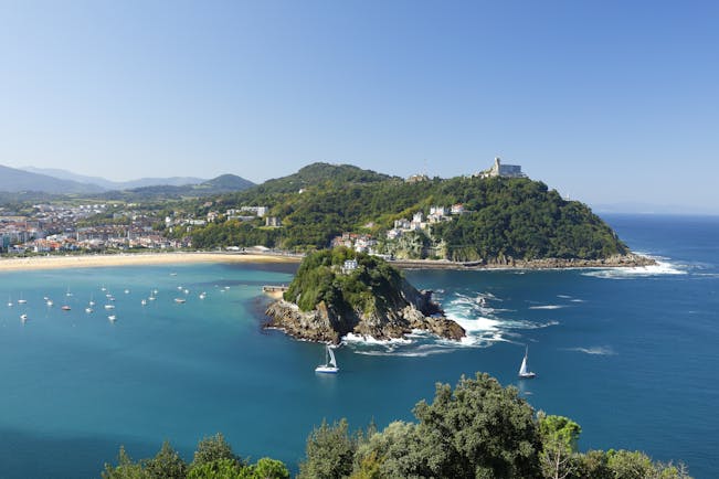 Sailing boats on blue sea in front of small island in bay with yellow sand beach in background at San Sebastian