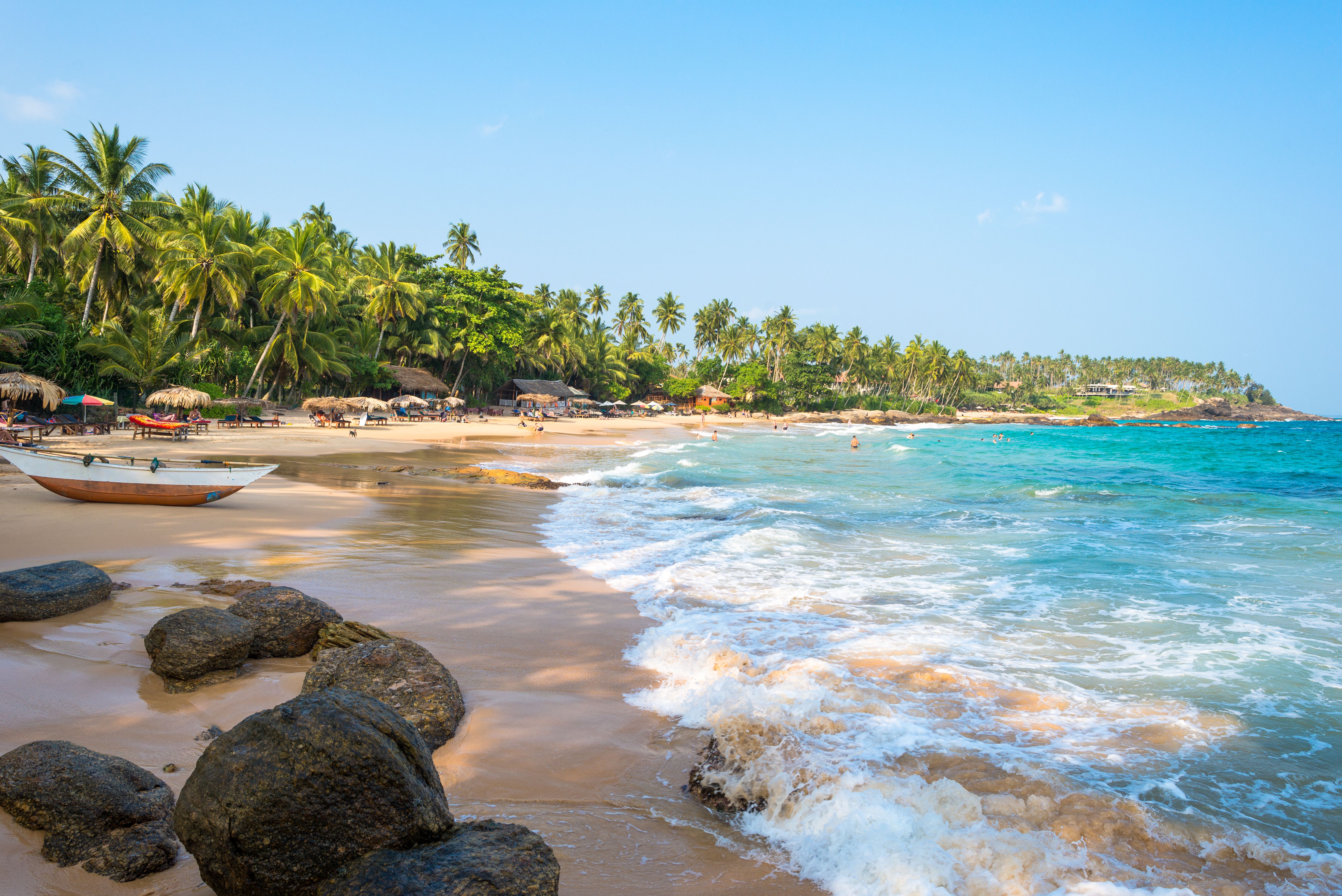 Beach in Tangalle, waves lapping golden sand, umbrellas and sun loungers, wooden boat, people swimming in the sea
