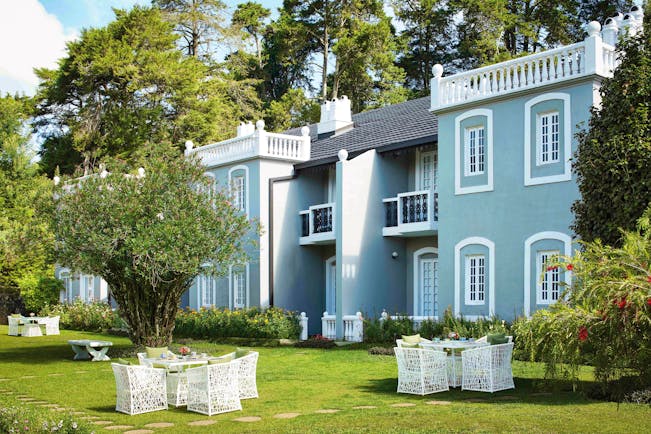 Hotel exterior showing a blue building with white lining, white chairs outside on the grass and trees