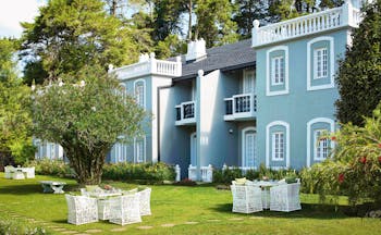 Hotel exterior showing a blue building with white lining, white chairs outside on the grass and trees