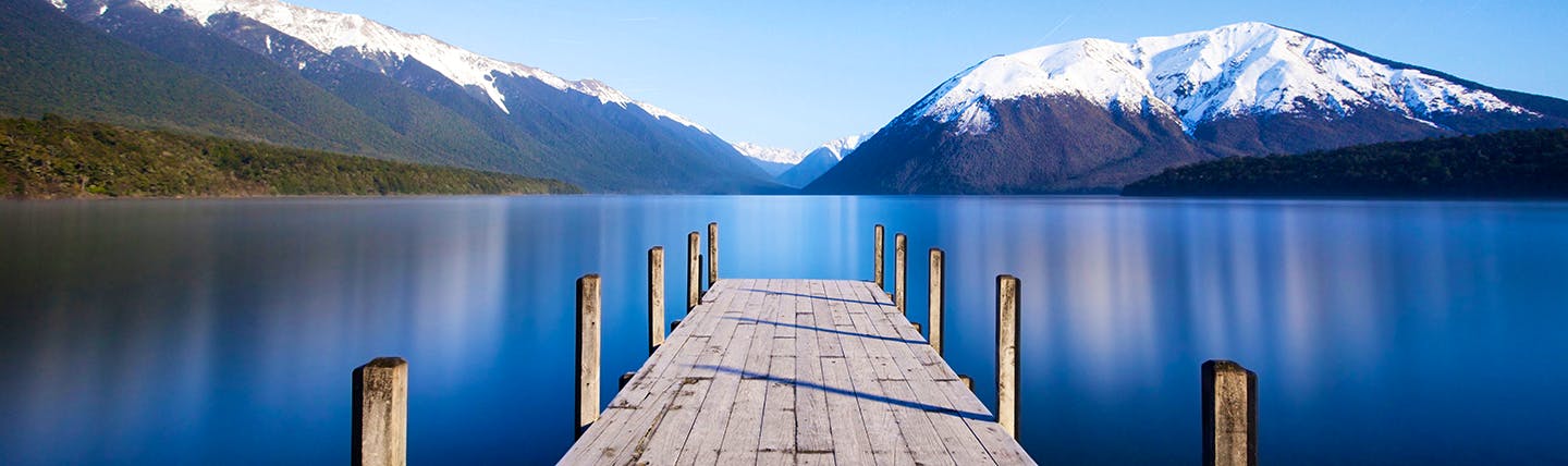 Wooden jetty into calm blue lake with snowy mountains in distance