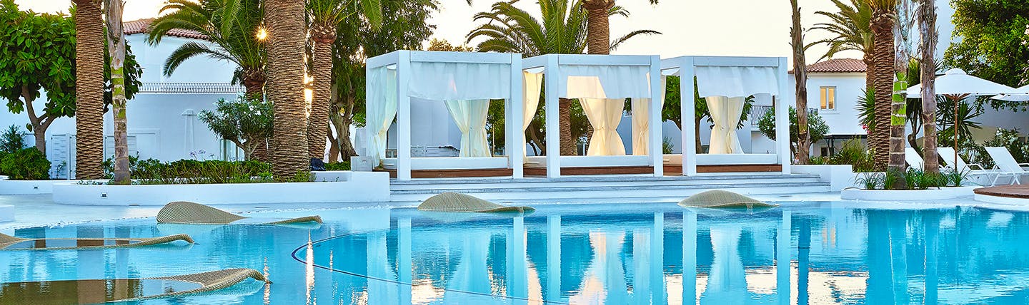 Blue swimming pool surrounded by large white canopy day beds and palm trees at Grecotel Caramel